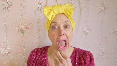 A person in a glittery pink dress and bright yellow hair bow looks into the camera applying bright pink lipstick. Behind them is a wall covered in pink flowery wall paper