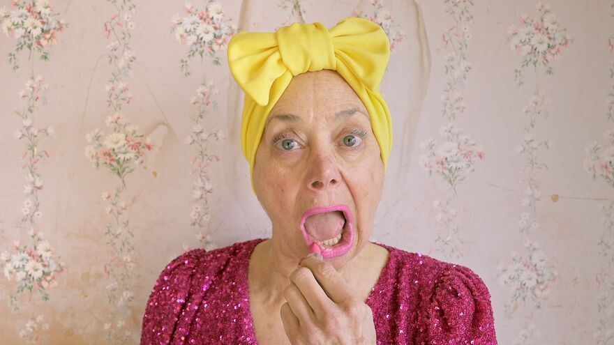 A person in a glittery pink dress and bright yellow hair bow looks into the camera applying bright pink lipstick. Behind them is a wall covered in pink flowery wall paper