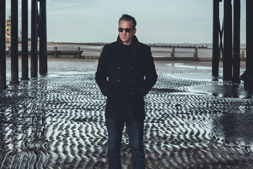 A white man wearing sunglasses, a black coat and jeans stands underneath a pier. The tide is out and you can see waves in the sand beneath him.