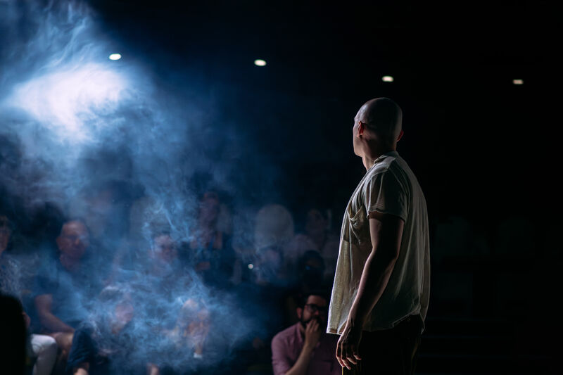 A white, bald performer stands with his back to the camera, looking out onto the audeicne, amidst haze and mood white lighting