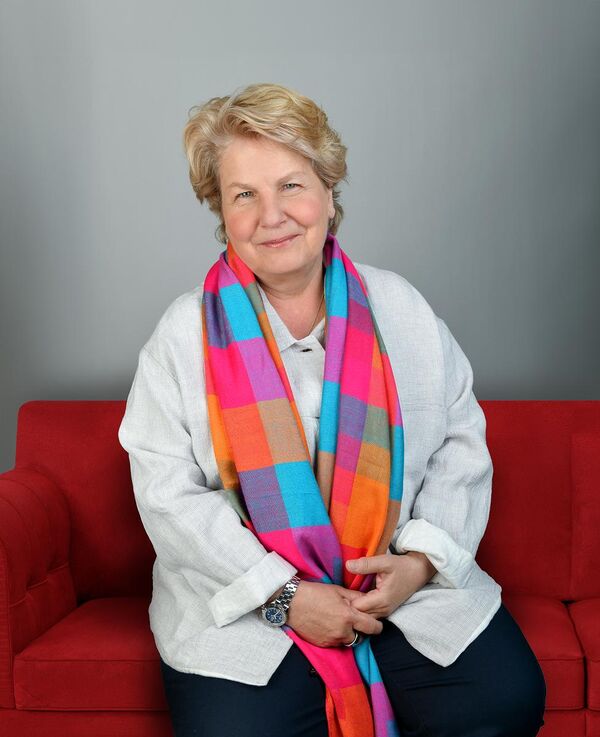 Sandi Toksvig smiling as she sits on a red couch wearing casual clothing and a colourful scarf
