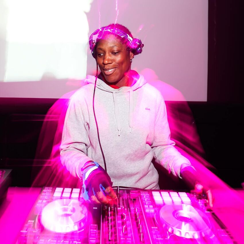 A DJ is spinning decks. They wear headphones and a light coloured jumper and have their hands on a set of DJ decks. There is a pink light which is seems to emit from them.
