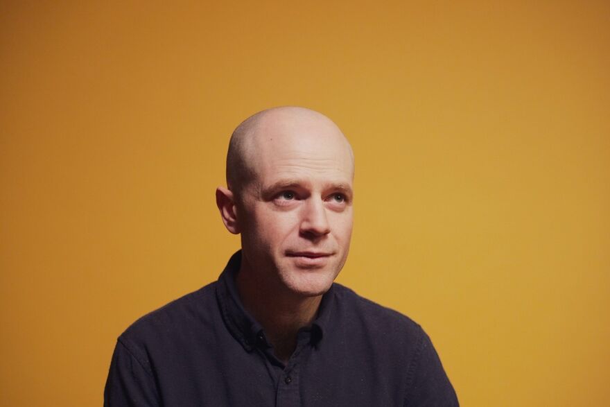 David Elms over a yellow background, he is wearing a dark blue shirt