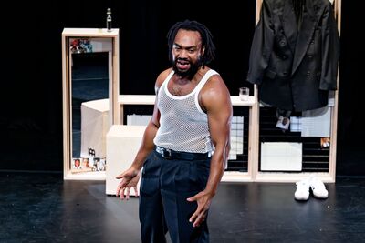 A man wearing a white vest standing on stage in front of wooden stage props and a mirror.