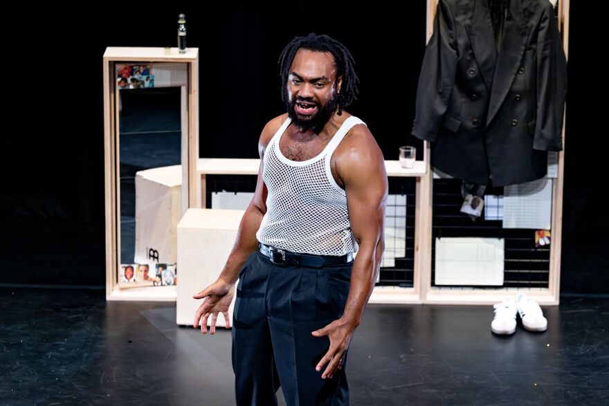 A man wearing a white vest standing on stage in front of wooden stage props and a mirror.