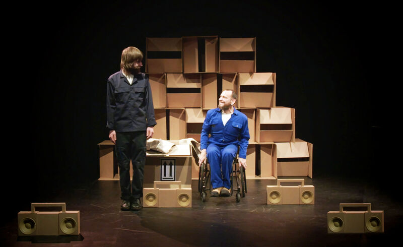 A man in a blue jumpsuit sits in his wheelchair in front of a wall of cardboard boxes. To his left stands another man in a dark blue jumpsuit. They are surrounded by 4 boomboxes made out of cardboard.