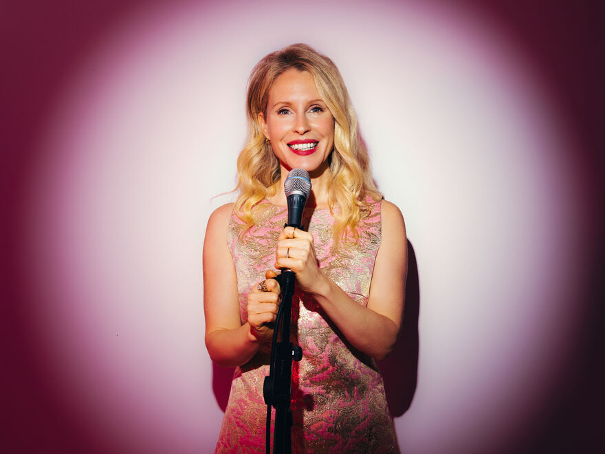 Lucy Beaumont smiling holding a microphone. She is standing in front of a white spotlight