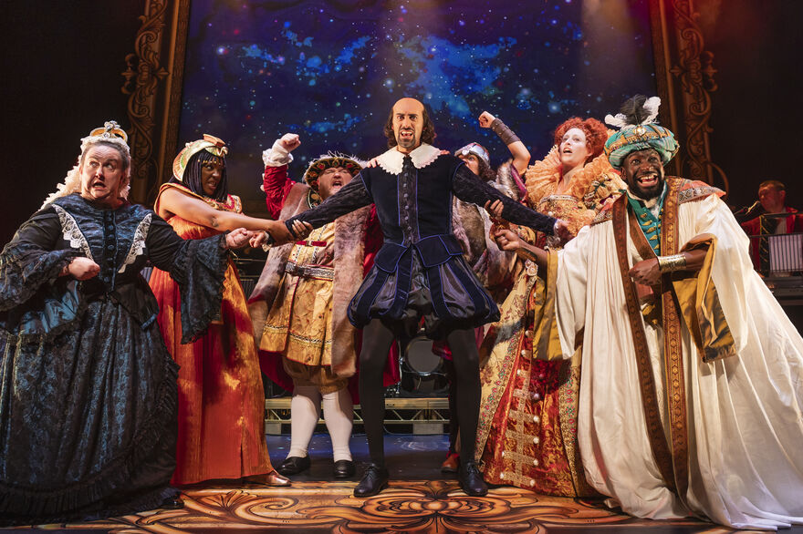 The cast of Horrible Histories are dressed as historical characters, surrounding William Shakespeare and pulling both his arms