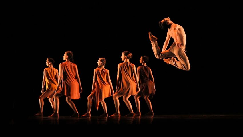 six people dance on a dark stage with the person foremost to the right jumps high up into the air