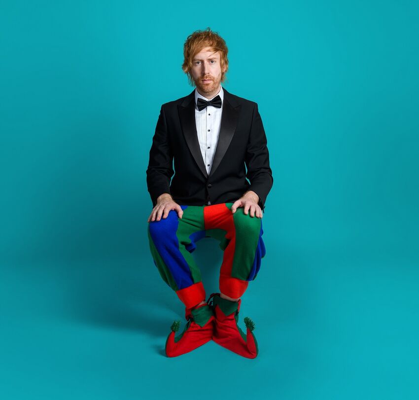 Mark Simmons, a white man with red hair and red beard, sits on a blue stool. He is wearing a black suit jacket and bow tie on top, and jester trousers and shoes on the bottom.