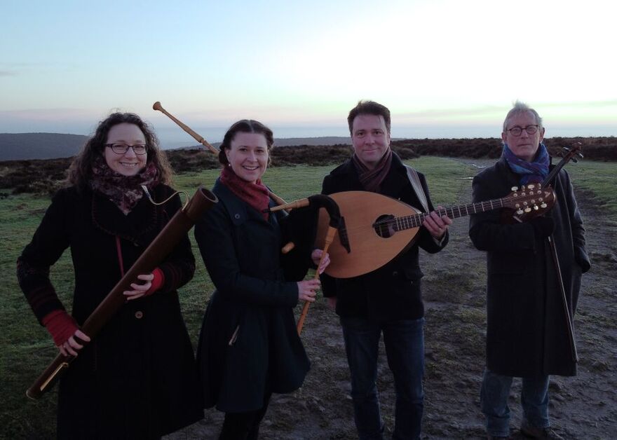 Four people standing on a field at dusk. They are smiling at the camera and hoping a selection of woodwind and string instruments. They are all wearing coats