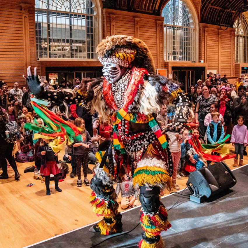 A dancer on stage in the Corn Exchange with his arm oustretched, wearing traditional African clothing. A crowd of smiling families stand in the background 