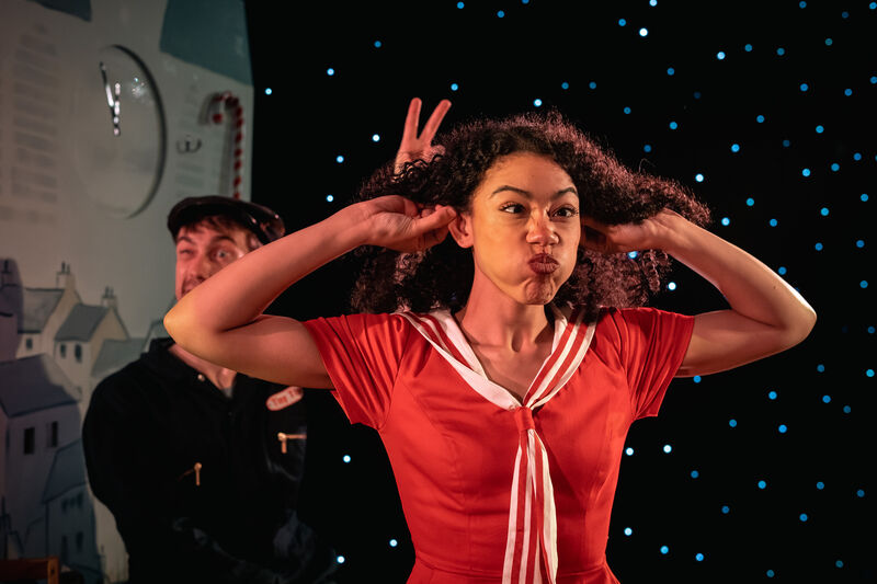 A girl in a red sailor dress holding her ears and pulls a silly face with a man holding two fingers behind her head in a teasing manner