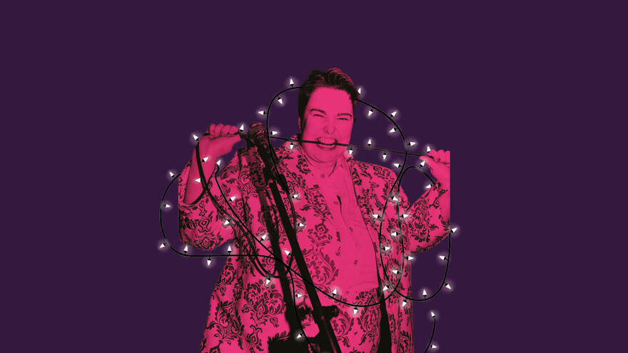 Erin Enfys is wearing a patterned suit and biting down on a microphone cable. The image has a pink wash, has a textured white outline and is overlayed on a purple background as if stuck on by hand. There are fairy lights wrapped around them