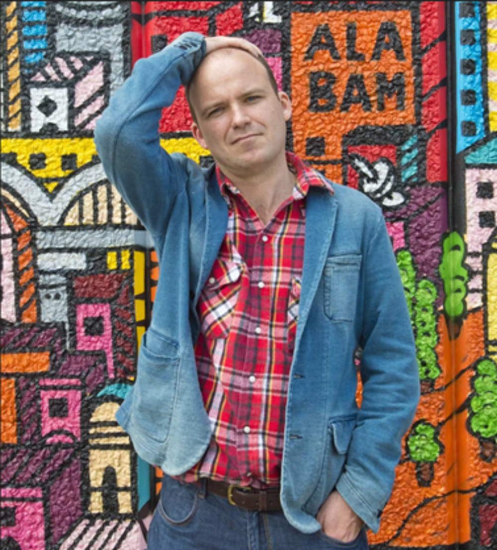 Rory Kinnear standing in front of a graffiti wall wearing a plaid shirt and blue jacket. He has one arm resting on his head