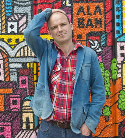 Rory Kinnear standing in front of a graffiti wall wearing a plaid shirt and blue jacket. He has one arm resting on his head