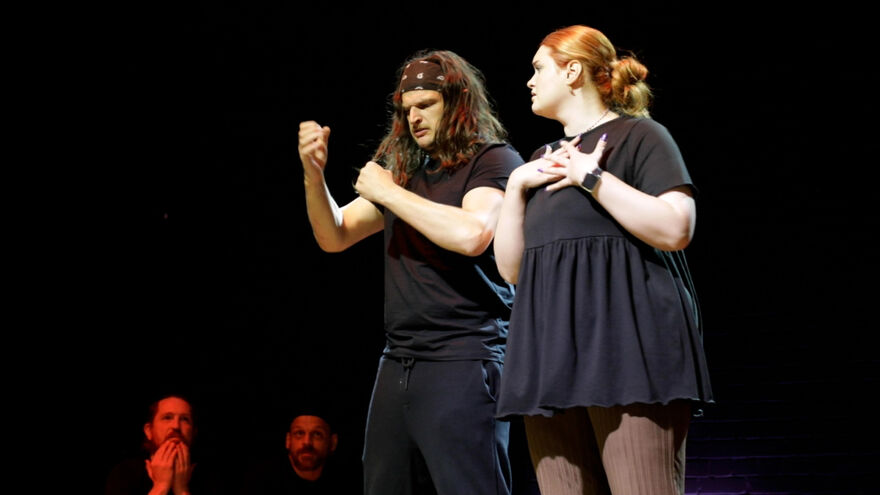 Two comedians stand onstage, lit by a single spotlight, mid-performance; one wears a black t-shirt and sweatpants with a bandana around their head keeping mid-length brunette hair off their face. The other wears a black dress and brown leggings, their ginger hair pulled back into a low bun. Two onlookers sitting on the floor to their left watch in amusement, lit in a soft red light.
