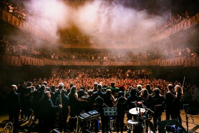 Big group od people standing on stage facing the crowd. It looks atmospheric with haze and orange lights