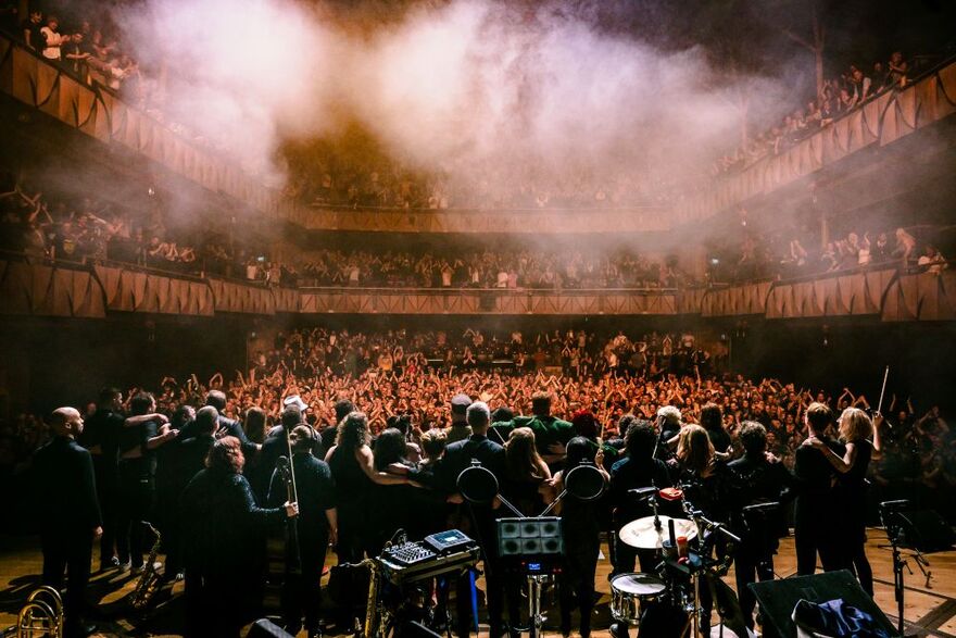 Big group od people standing on stage facing the crowd. It looks atmospheric with haze and orange lights